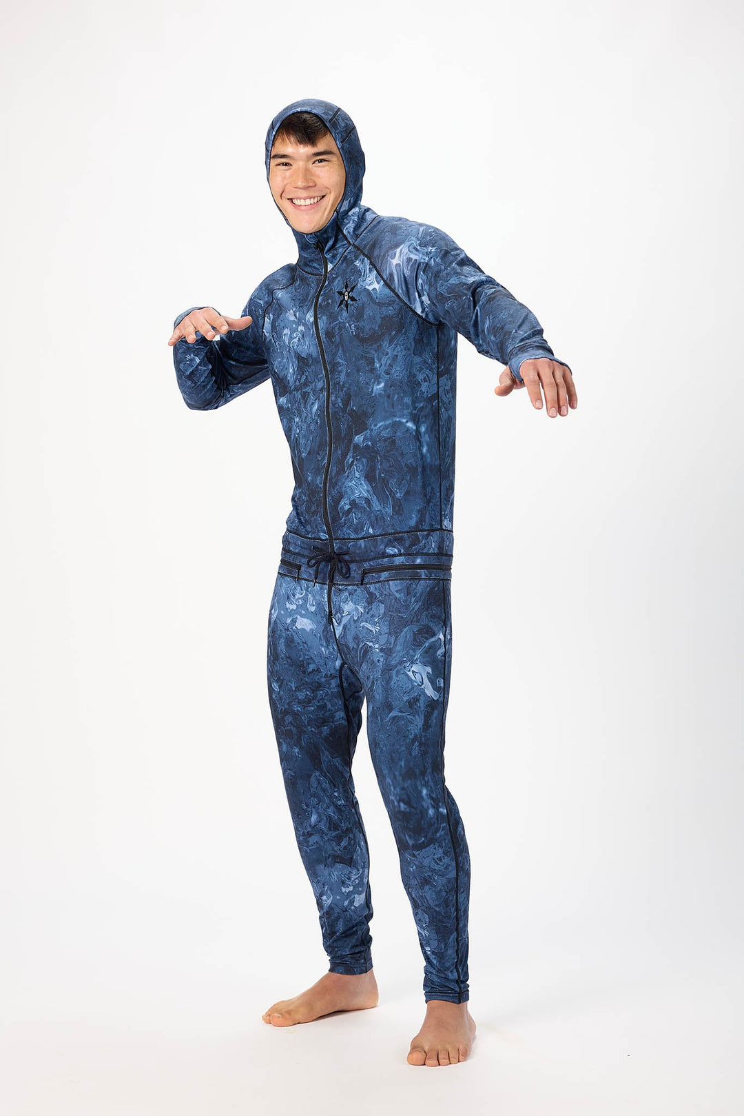 A man wearing a blue swirl patterned onesie with a hooded with a zipper and jogger pants, standing against a plain white studio background.