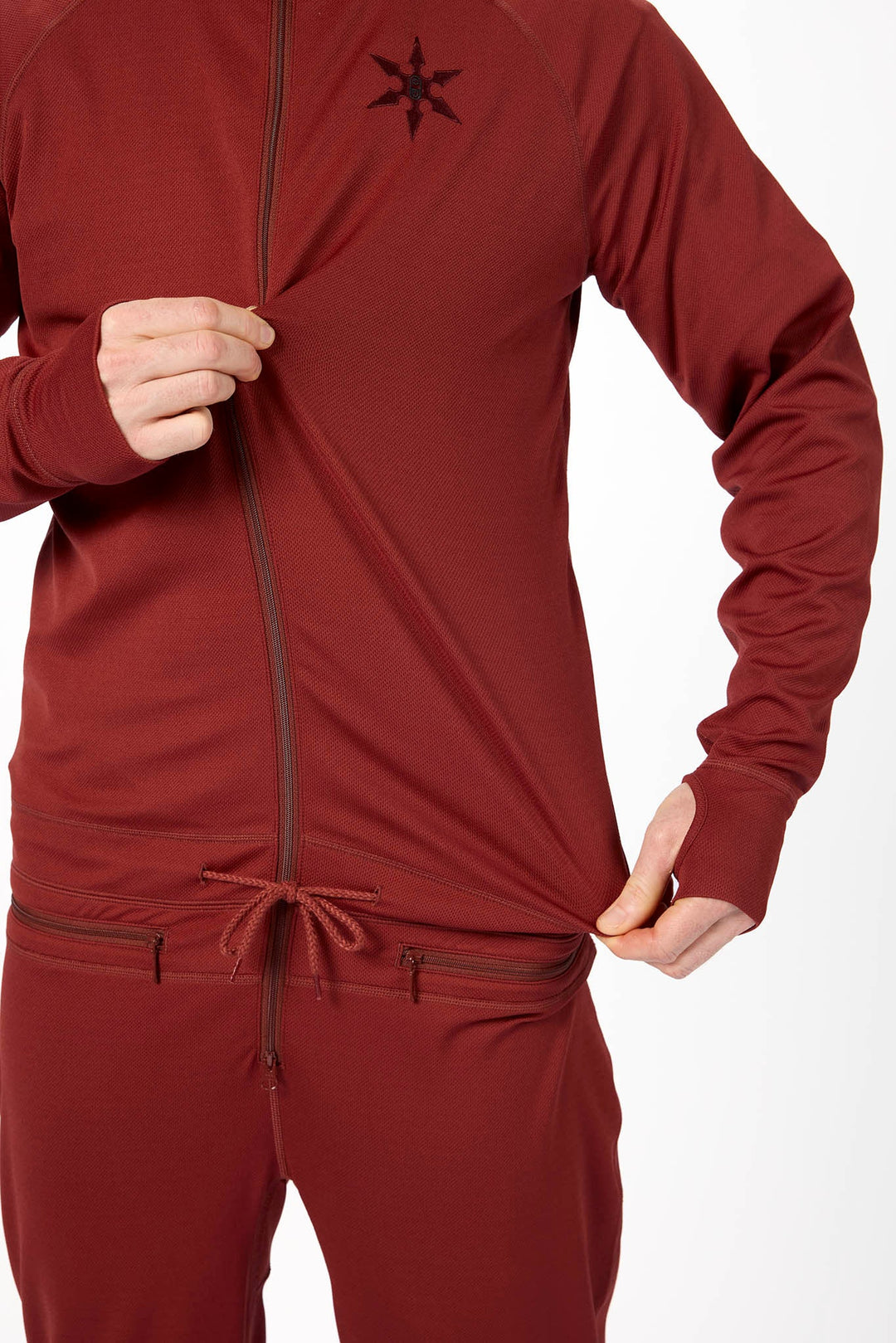 A close-up of a person wearing a matching burgundy zip-up athletic jacket and jogger pants, pulling the jacket hem outward with both hands. 
