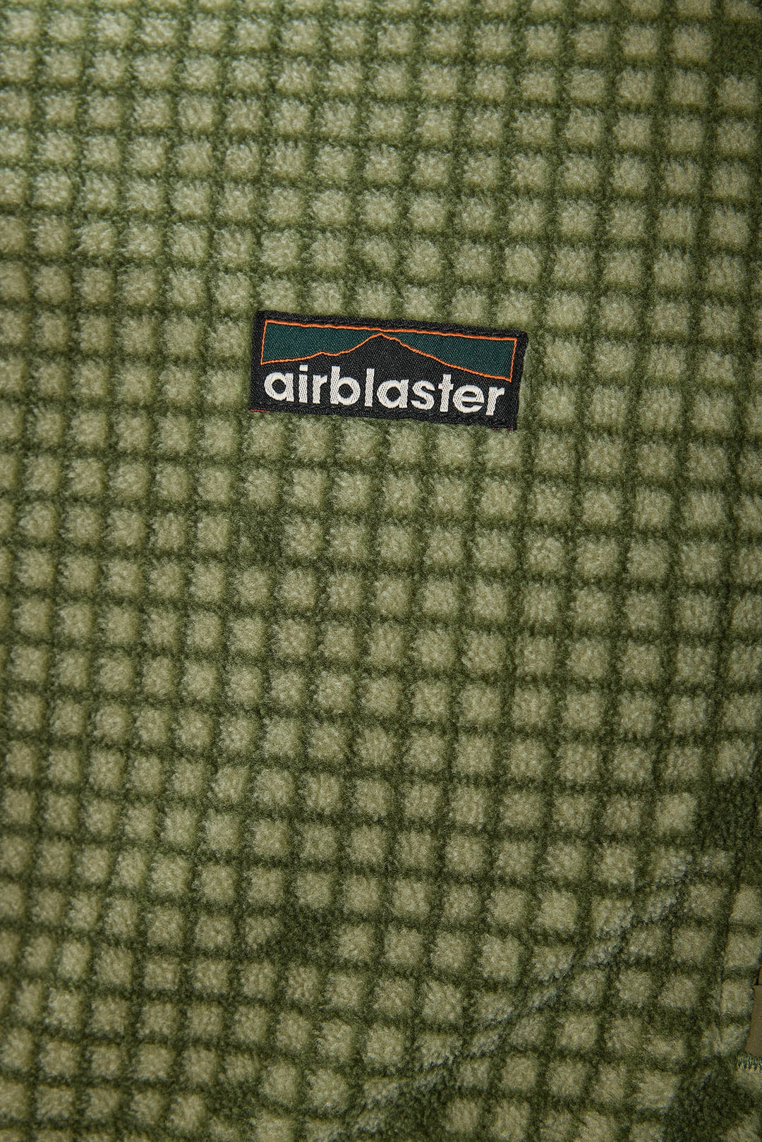 Close-up of a textured fleece fabric with a green and beige grid pattern, featuring a central black Airblaster logo patch.