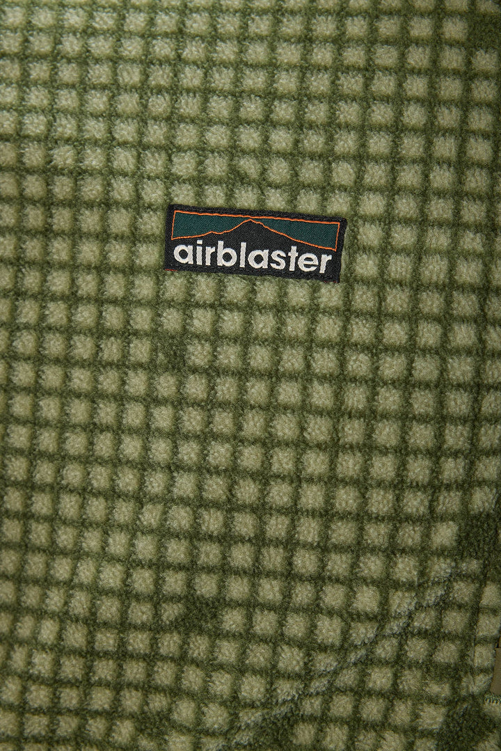 Close-up of a textured fleece fabric with a green and beige grid pattern, featuring a central black Airblaster logo patch.