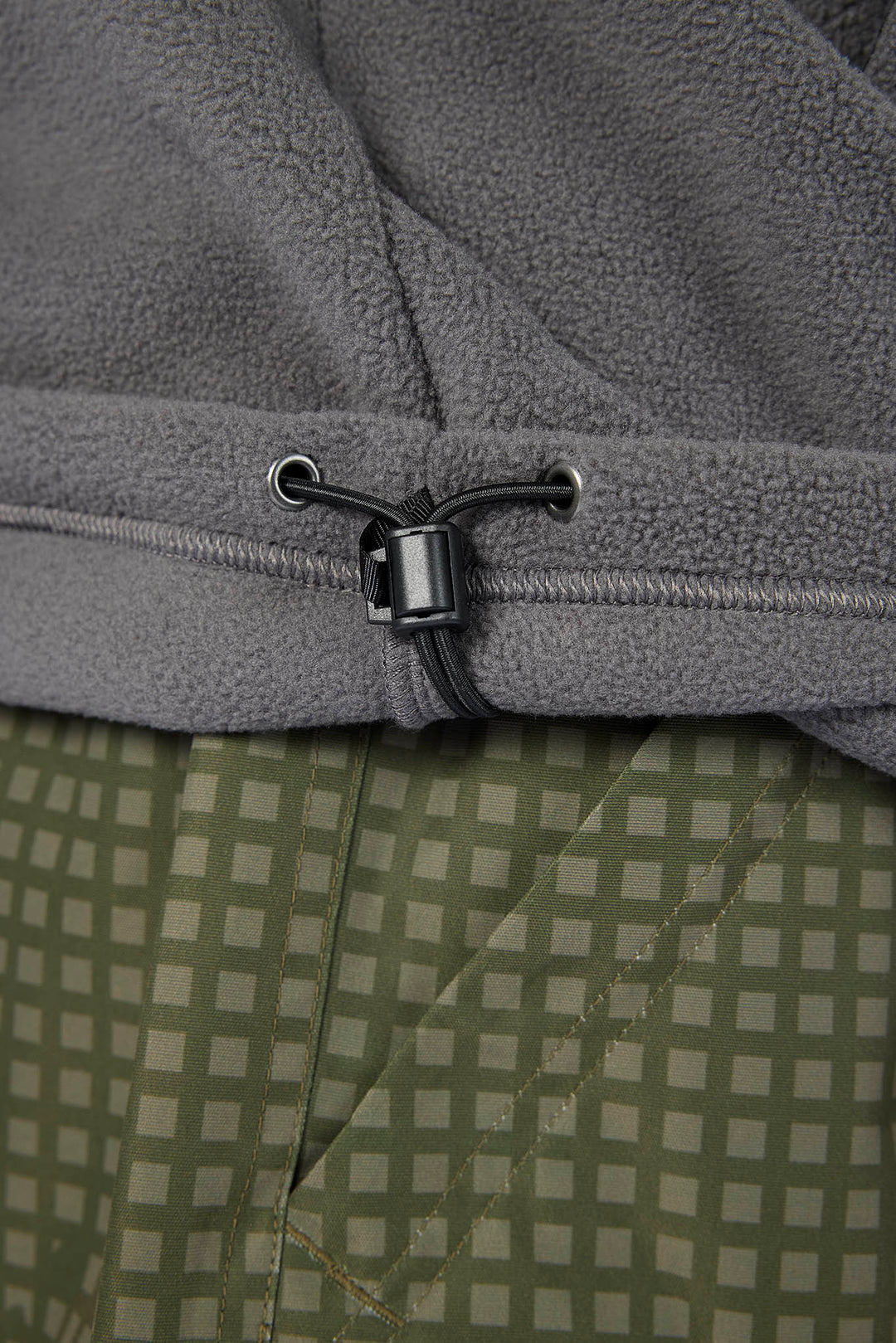 Close-up of a gray fleece garment with a black drawstring toggle, layered over an olive green and beige checkered fabric.