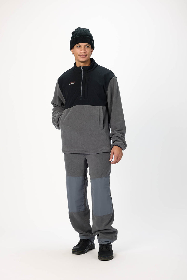 A man modeling a two-tone gray and black fleece quarter-zip pullover and matching gray pants with black shoes and a black beanie, photographed against a plain white studio background.