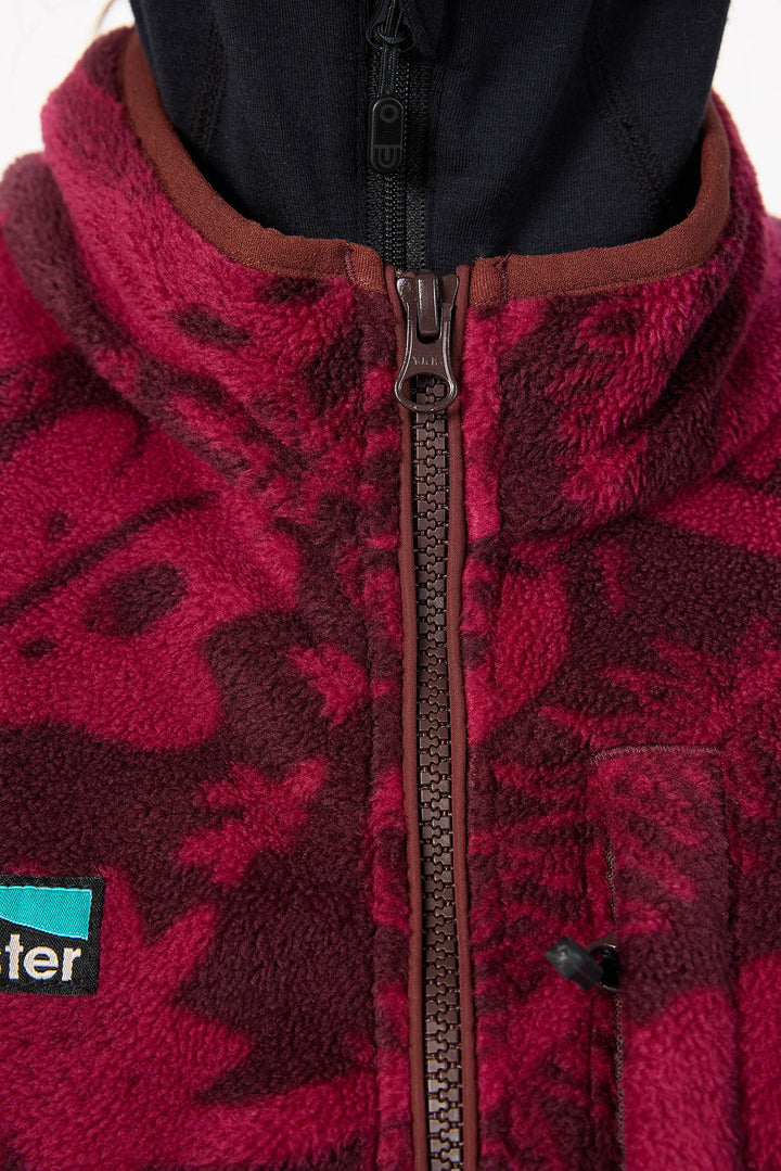 Close-up of a fleece jacket with a dark red and black camouflage pattern, a brown collar, and a black zipper. A small black and teal Patagonia logo patch is visible on the lower left.