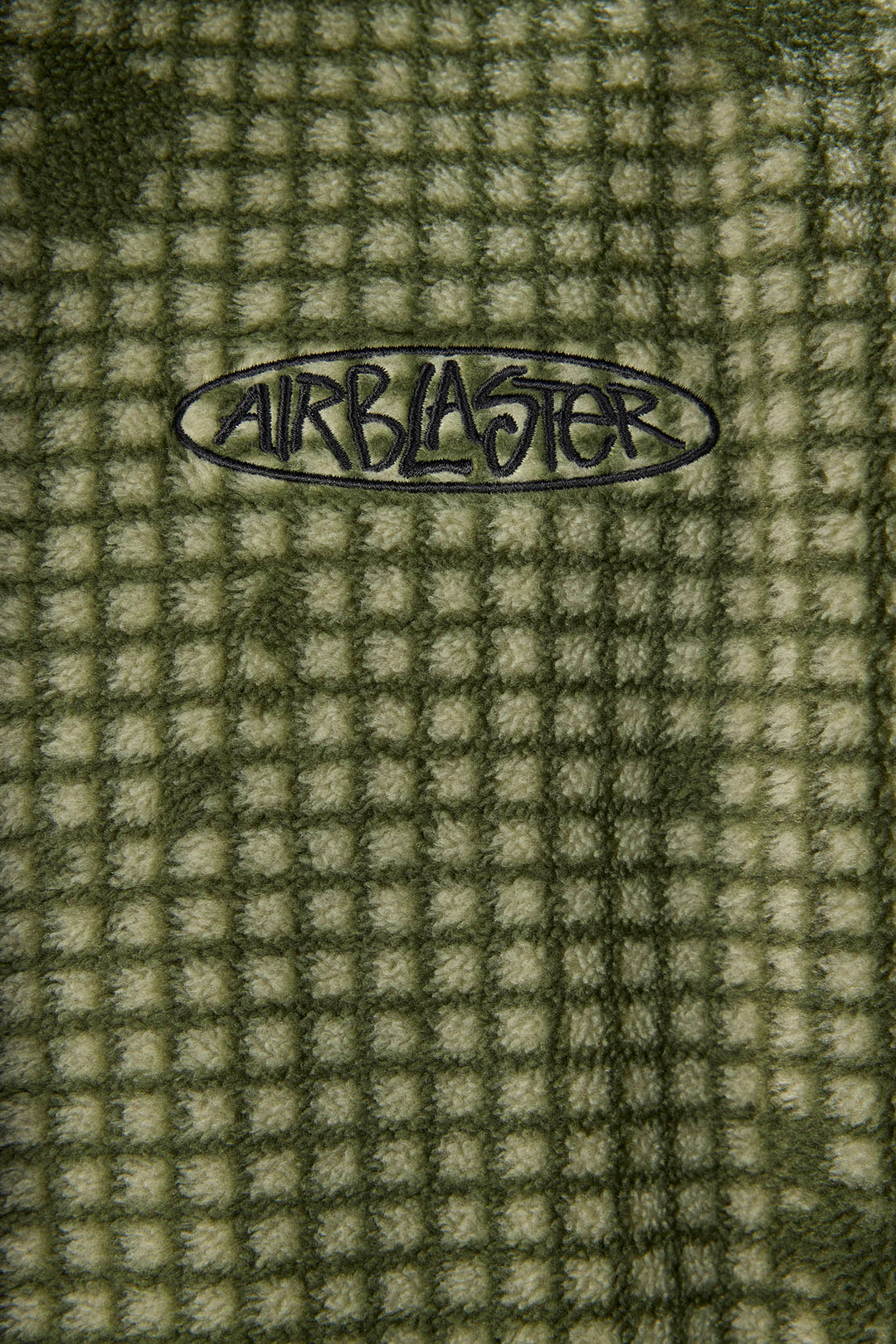 Close-up of a textured waffle fleece blanket in olive green and beige, featuring a black Airblaster logo embroidered in the center.