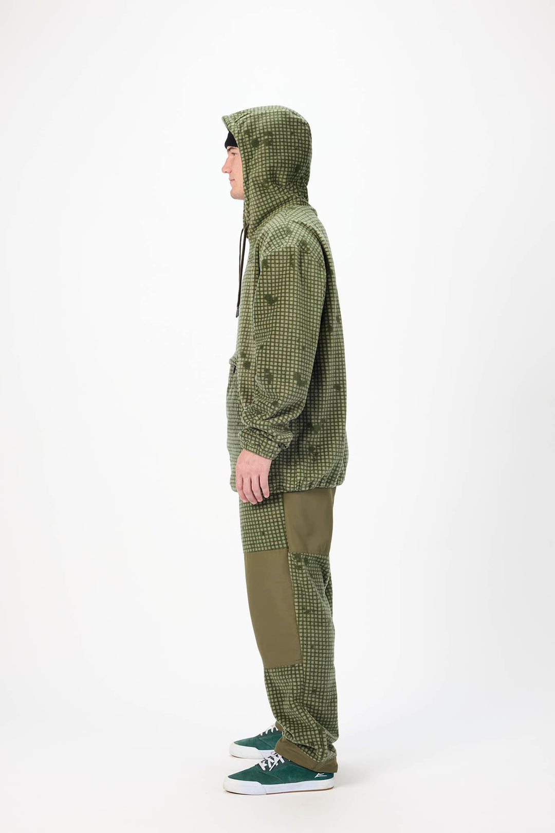 A person shown in side profile wearing an olive green hooded jacket with a subtle tonal pattern, matching olive pants, and green sneakers against a plain white studio background.