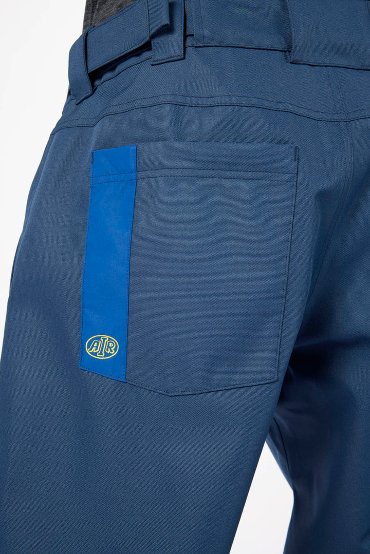 Close-up of the back waistband and right pocket of blue work pants, featuring a vertical blue stripe on the pocket with a small circular logo patch.