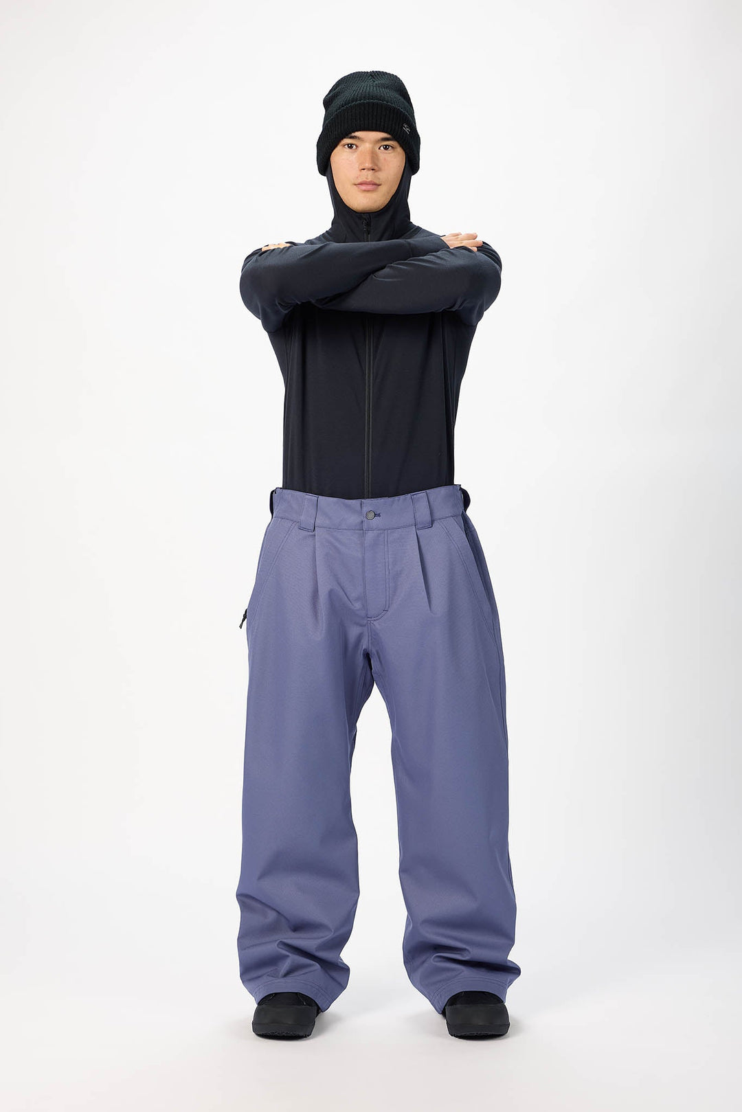 A model wearing a black zip-up jacket, a black beanie, and slate blue snow pants, standing with arms crossed against a white studio background.