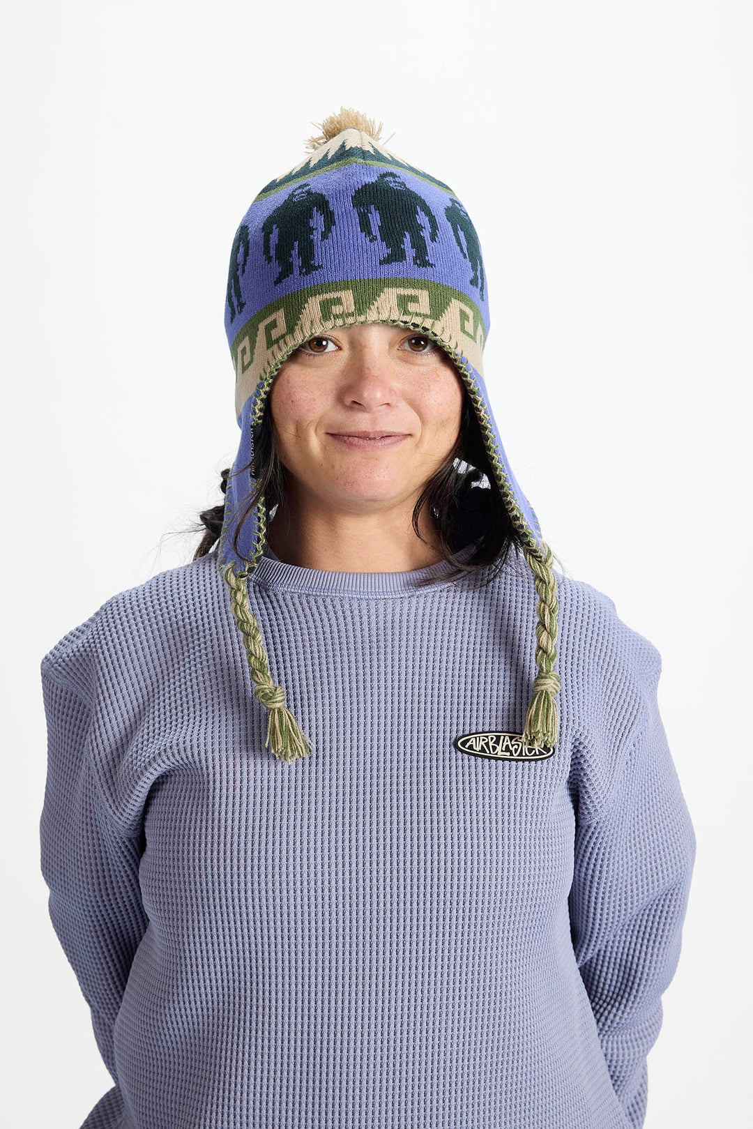 A woman wearing a purple knit winter hat with ear flaps, braided ties, and a pom-pom, paired with a matching purple waffle-knit sweatshirt. The hat features black silhouettes and a patterned band.