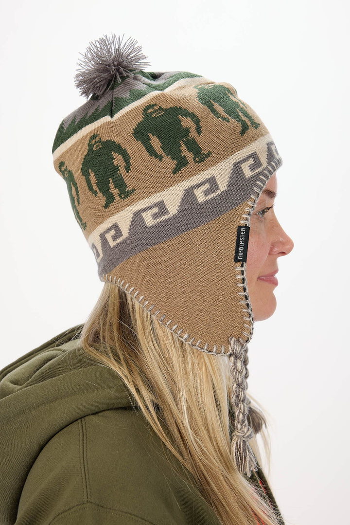 A woman in profile wearing a knit winter hat with a gray pom-pom, green gorilla patterns, and ear flaps, against a plain white background.