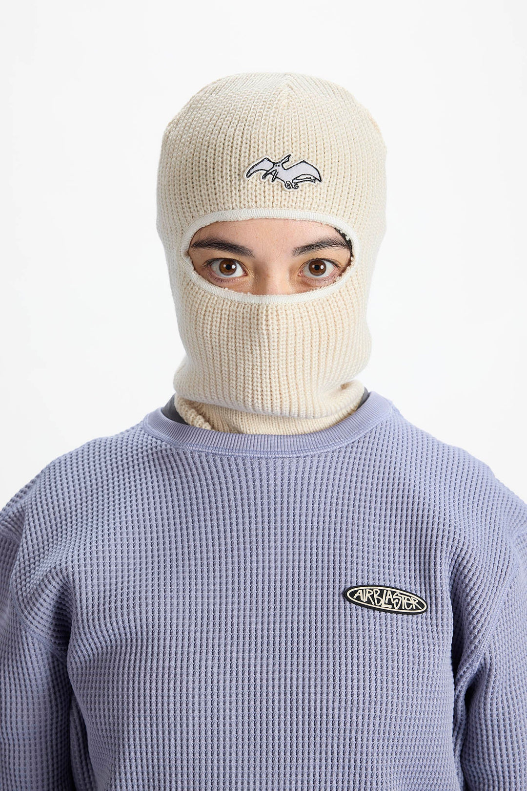 A model wearing a cream-colored ribbed knit balaclava mask with a small embroidered logo, paired with a lavender waffle-knit sweatshirt. The background is a clean, solid white studio backdrop.
