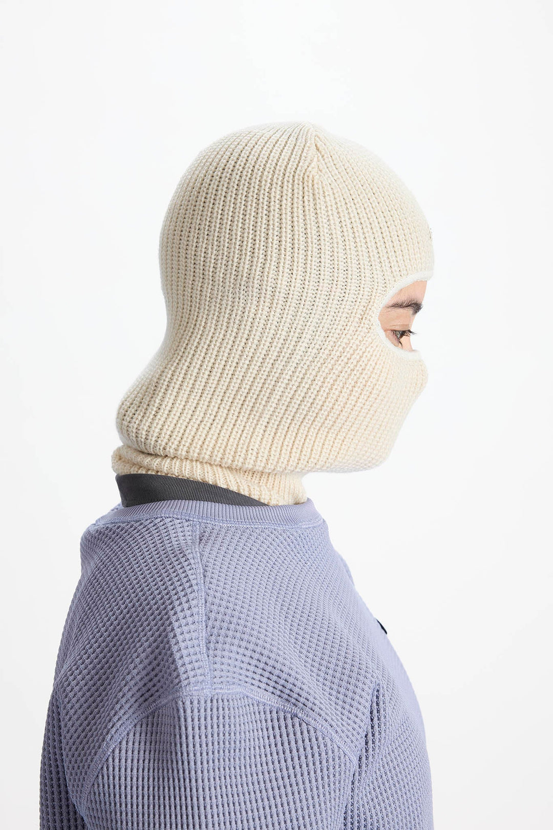 A side profile view of a person wearing a cream-colored ribbed knit balaclava mask and a lavender waffle-knit top against a plain white background.
