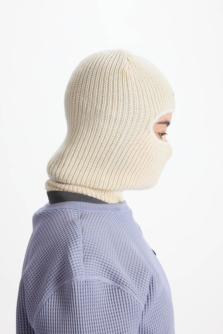 A side profile view of a person wearing a cream-colored ribbed knit balaclava mask and a lavender waffle-knit top against a plain white background.