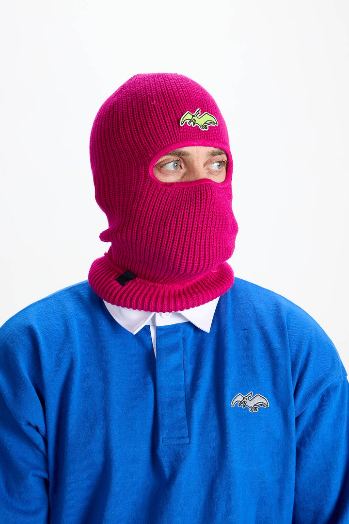 A person wearing a bright magenta knit balaclava mask with a small embroidered logo on the forehead, paired with a blue polo shirt against a plain white background.