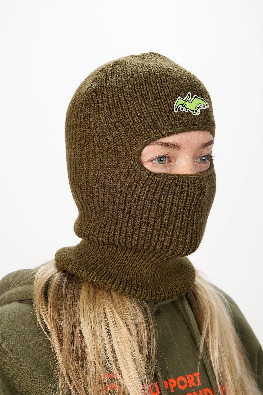 A person wearing an olive green ribbed knit balaclava mask with a small green and yellow embroidered logo on the forehead, shown against a plain light background.
