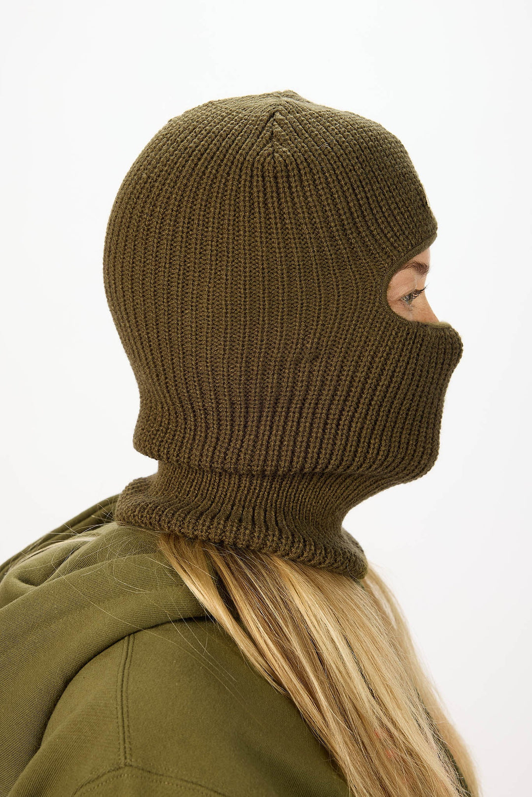 A person shown in side profile wearing an olive green ribbed knit balaclava mask and matching hooded jacket against a plain white background.