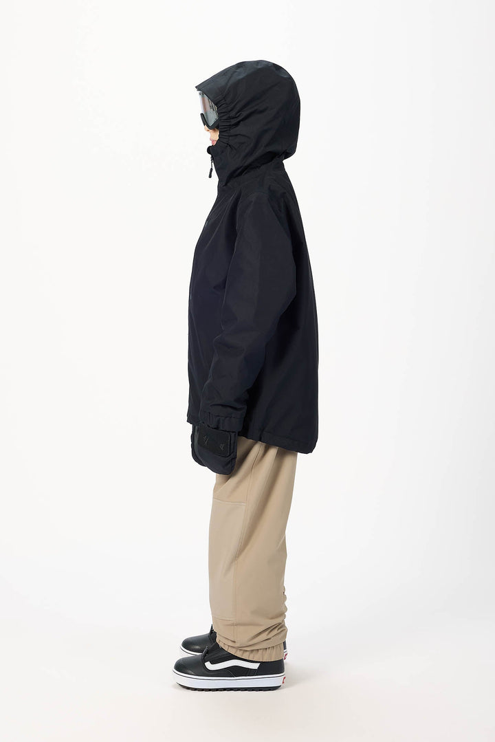 A person shown in profile wearing a black hooded jacket, beige pants, and black sneakers against a plain light gray studio background.