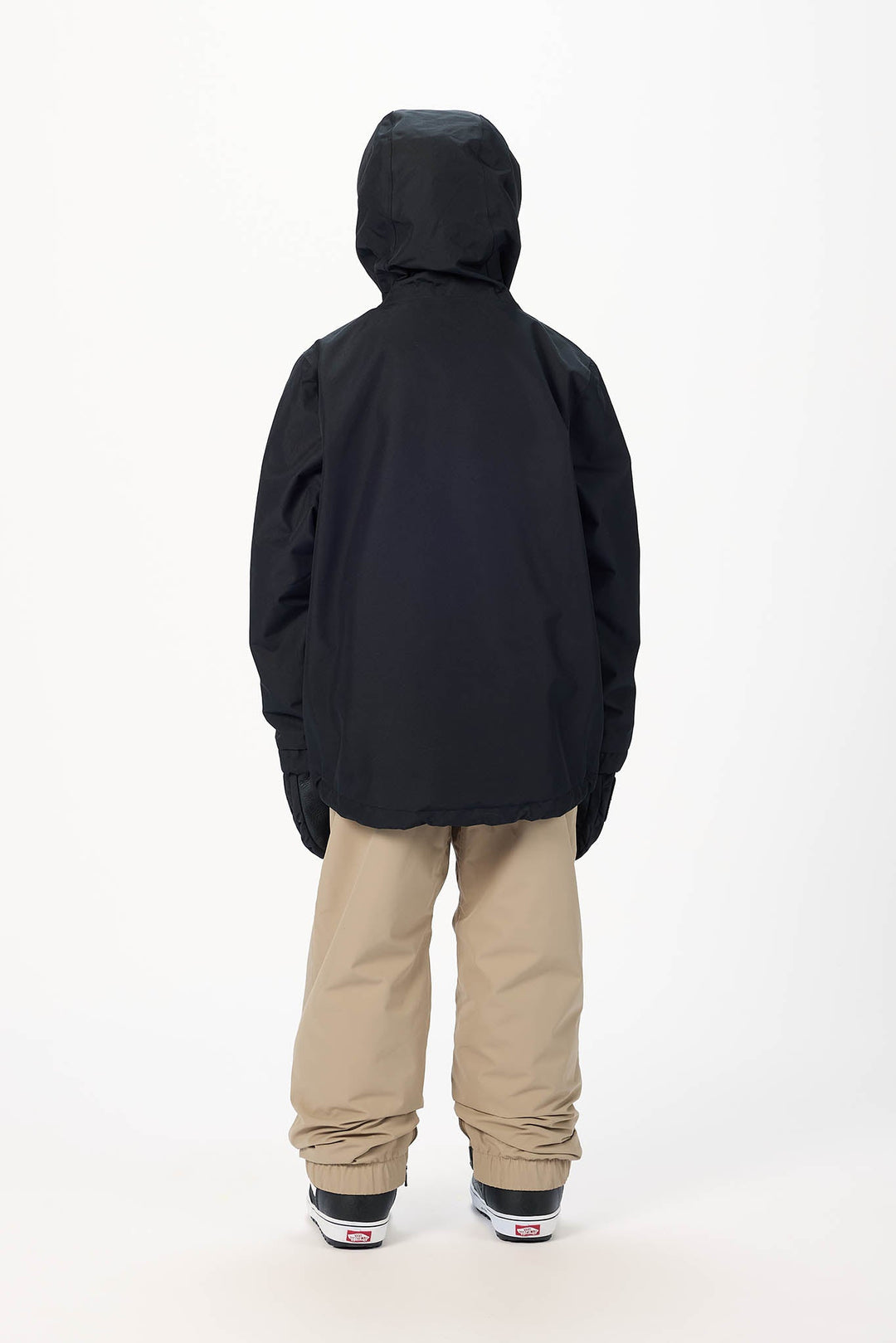 A child viewed from the back wearing a black hooded jacket, beige snow pants, and black sneakers with white soles, standing against a plain white studio background.