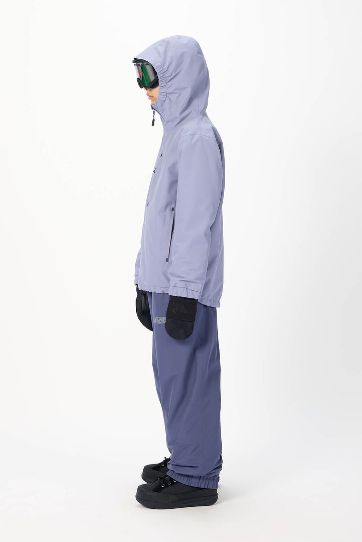 A child model shown in profile wearing a light periwinkle hooded jacket, matching pants, black gloves, and black snow boots, with ski goggles resting on the hood, against a plain white studio background.