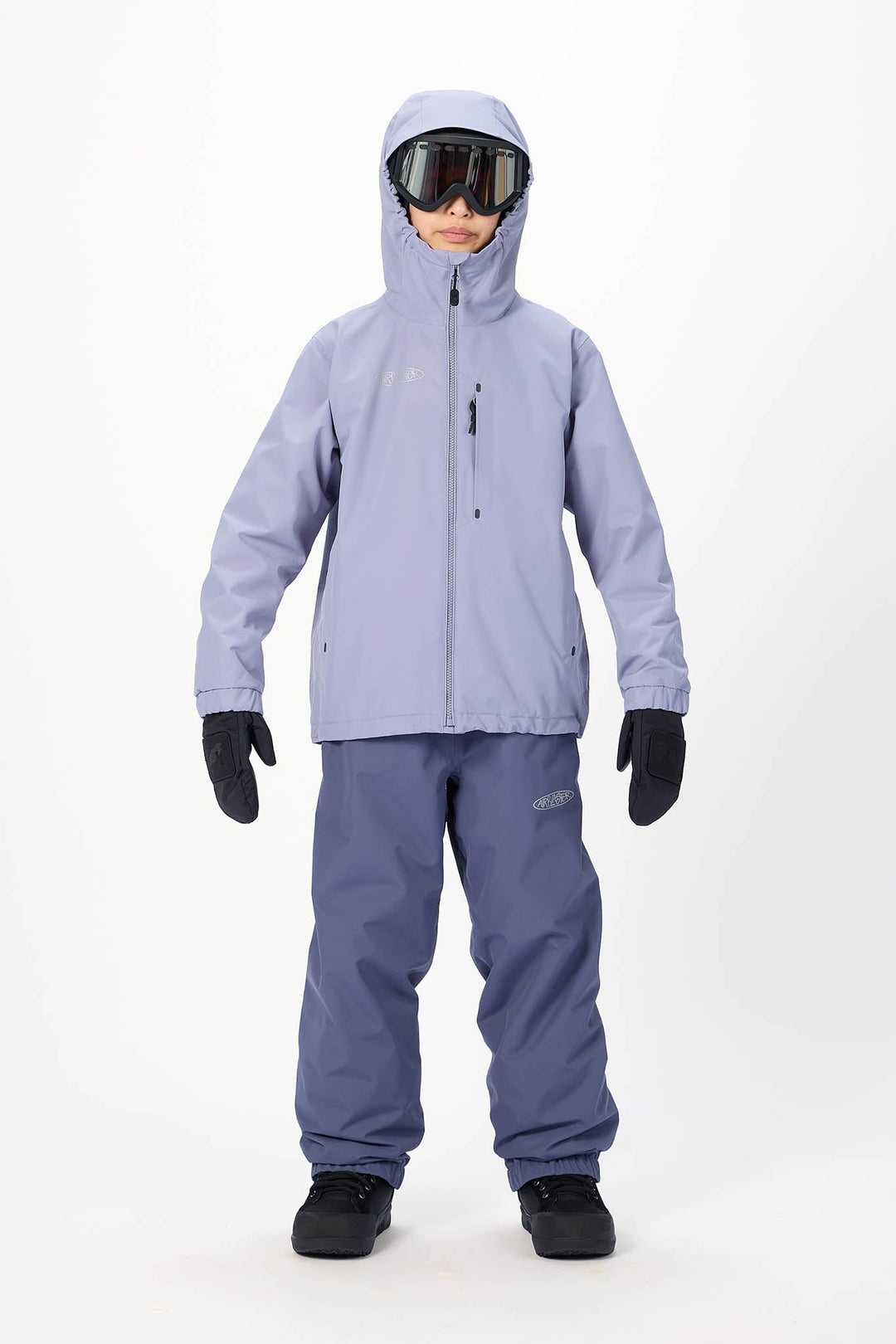 A child model wearing a light lavender ski jacket, matching snow pants, black gloves, and goggles, standing against a plain white studio background.