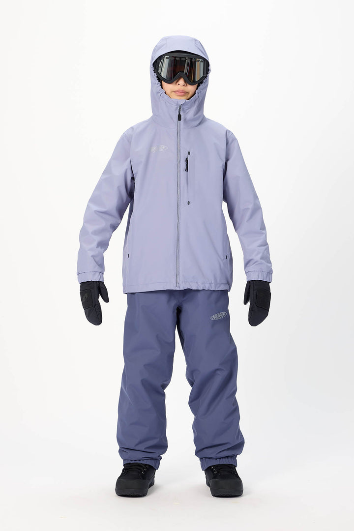 A child model wearing a light lavender ski jacket, matching snow pants, black gloves, and goggles, standing against a plain white studio background.