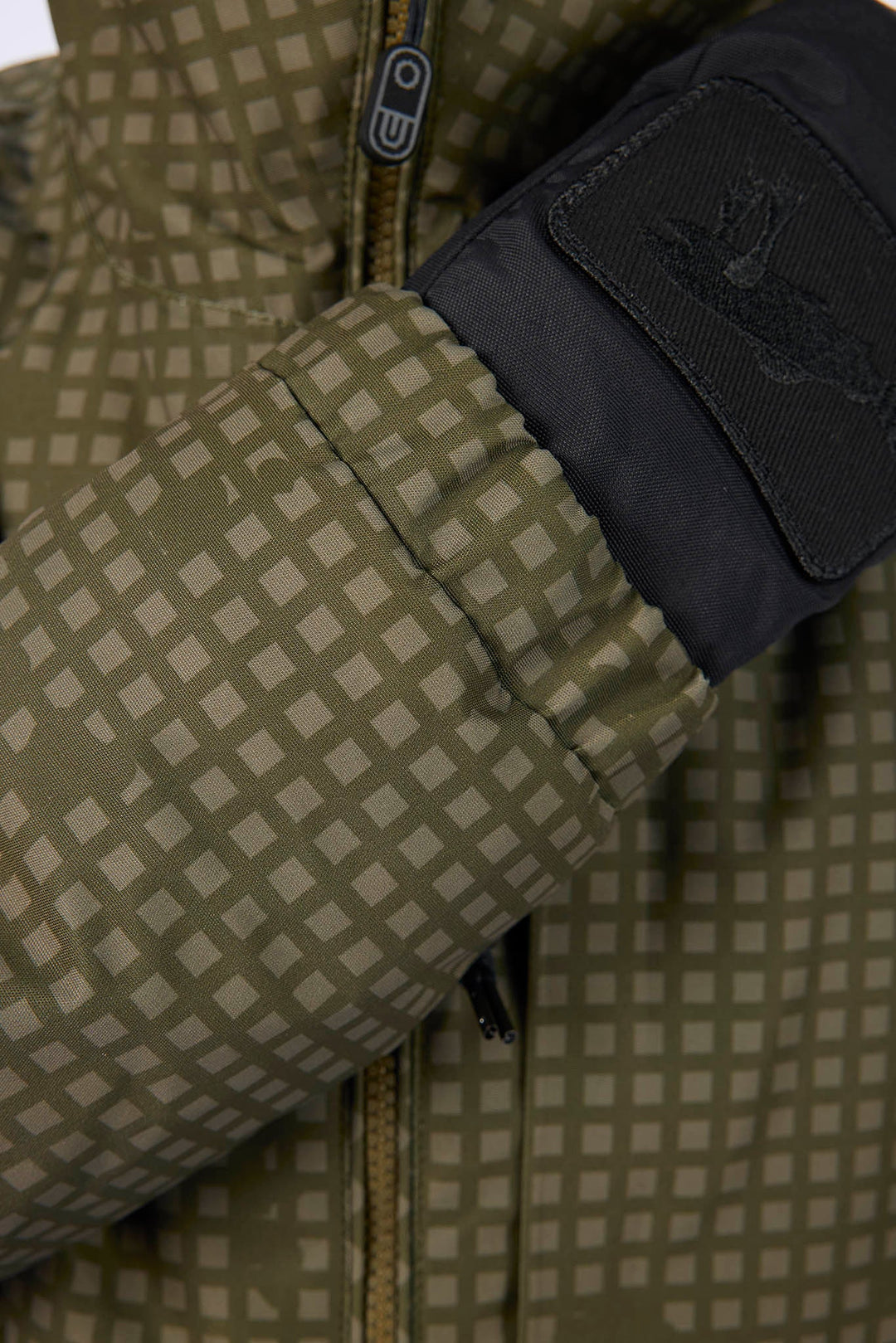 Close-up of a puffer jacket sleeve with a beige and olive green checkered pattern, a black cuff, and a black patch featuring a stylized logo.