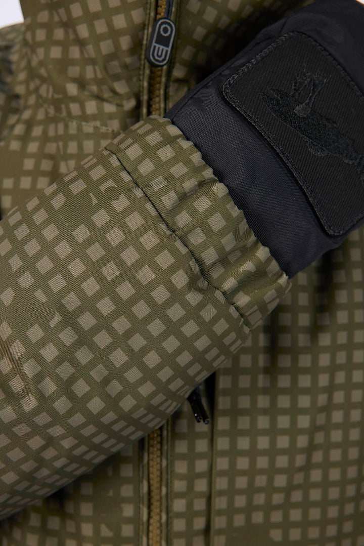 Close-up of a puffer jacket sleeve with a beige and olive green checkered pattern, a black cuff, and a black patch featuring a stylized logo.