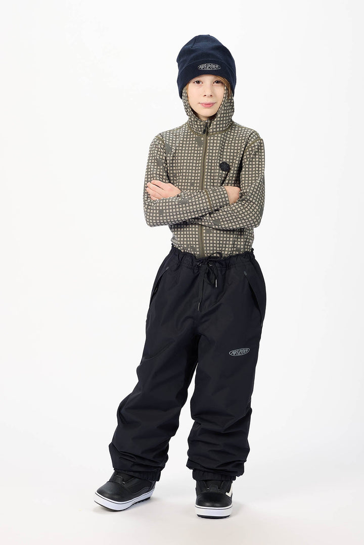 A child model wearing a patterned zip-up jacket, black snow pants, a dark navy beanie, and black sneakers, standing against a plain white studio background.