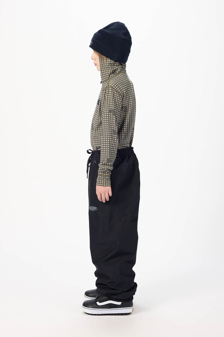 A child model shown in profile wearing a dark navy knit beanie, a green and beige patterned long-sleeve top, black drawstring sweatpants, and black sneakers with white soles, photographed against a clean white studio background.