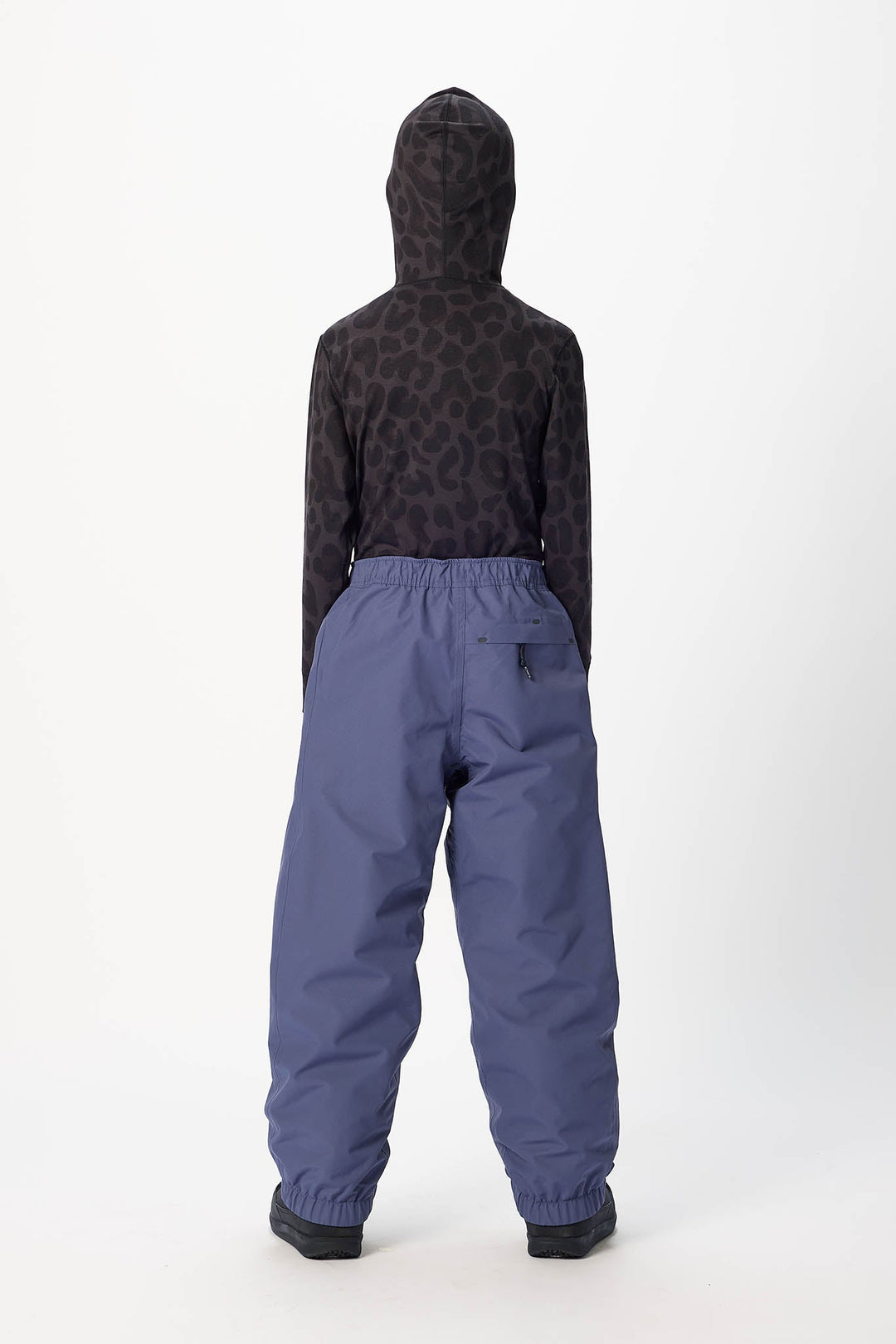 A child shown from the back wearing a dark patterned hooded sweatshirt and slate blue snow pants against a plain white studio background.