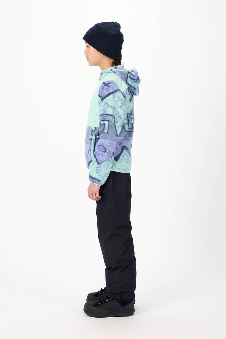 A child standing in profile wearing a light blue and purple patterned hoodie, black pants, black shoes, and a black beanie against a plain white studio background.