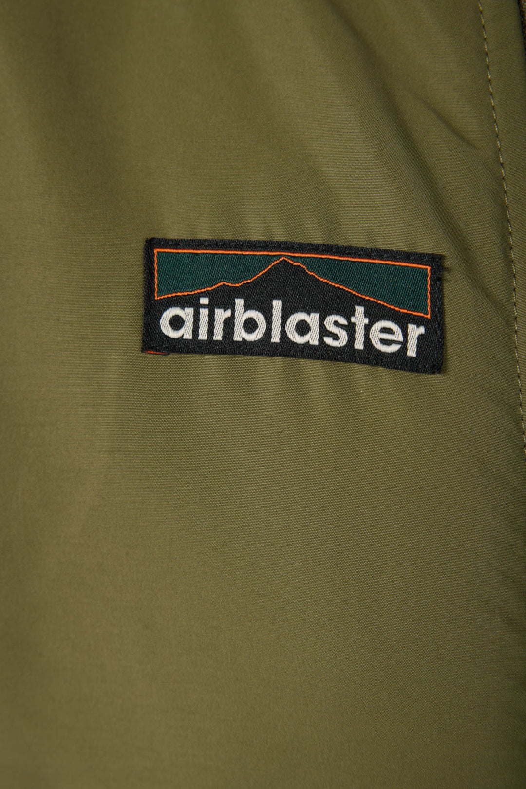 Close-up of an olive green jacket or fabric panel featuring a rectangular black Airblaster logo patch with white text and a dark teal and orange graphic.