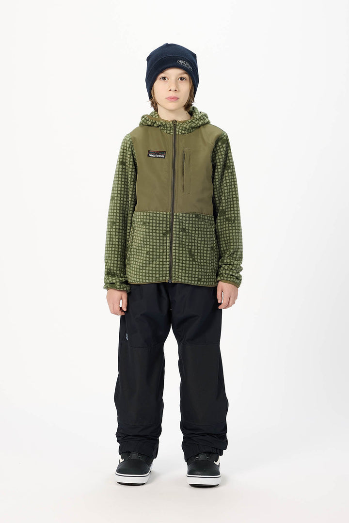 A child model wearing a green and black patterned hooded jacket, black snow pants, a dark navy beanie, and black sneakers, standing against a plain white studio background.